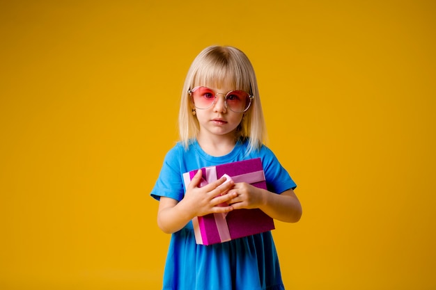 Kindermädchenblondine lächelnd in einem blauen Kleid, das eine rosa Geschenkbox auf einem gelben Hintergrundisolat hält