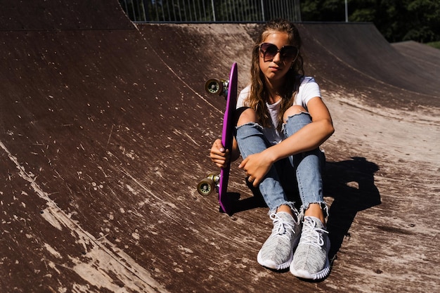 Kindermodell mit Brille sitzt und posiert mit Penny Board auf dem Skate-Spielplatz Aktives Kindermädchen mit extremem Lebensstil auf der Sportrampe