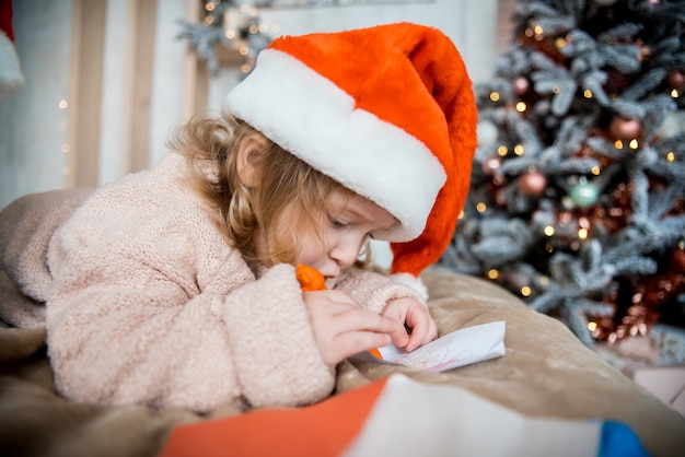 Kinderschreibensbrief zu Santa Claus-Brief im roten Hut. Familie an Weihnachten. Helles Wohnzimmer mit komin am Silvesterabend Kuscheliger warmer Winterabend zu Hause