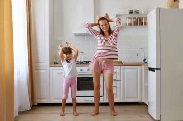 Kindertochter und Mutter, die sich zu Hause in der Küche amüsieren, Leute, die T-Shirts und Shorts tragen, zusammen tanzen, Hasenohren machen, in die Kamera schauen, haben positive Ausdrücke.