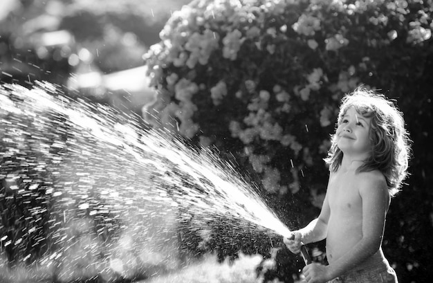 Kindesjunge, der die Pflanzen mit einem Wasserschlauch im Garten im Hinterhof des Hauses bewässert.