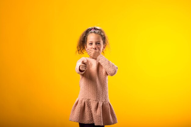 Kindesmädchen, das jemanden verspottet und neckt, zeigt den Finger in die Kamera und schließt den Mund über gelben Hintergrund. Bulling-Konzept