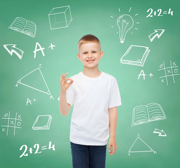 Kindheit, Geste, Bildung, Werbung und People-Konzept - lächelnder Junge im weißen T-Shirt mit OK-Schild über grünem Brett mit Kritzeleienhintergrund