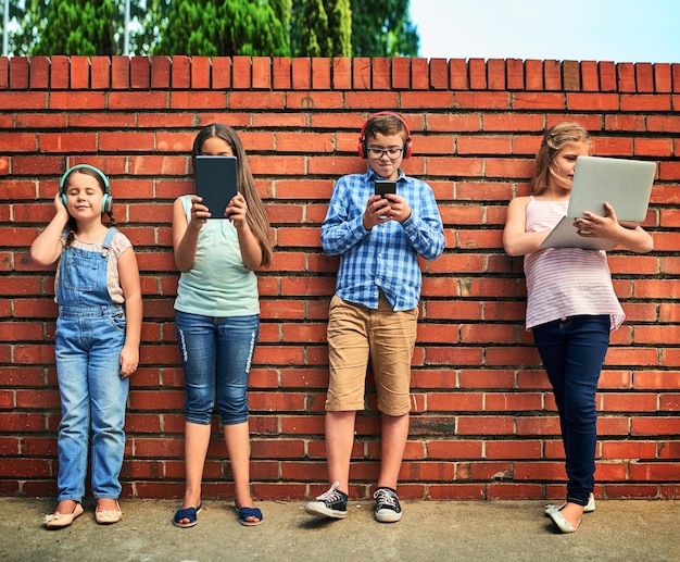 Kindheit im digitalen Zeitalter Aufnahme einer Gruppe kleiner Kinder, die digitale Geräte vor einer Ziegelwand verwenden