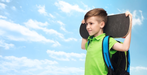 Kindheits-, Freizeit-, Schul- und Personenkonzept - glücklicher lächelnder Studentenjunge mit Rucksack und Skateboard über blauem Himmel und Wolkenhintergrund