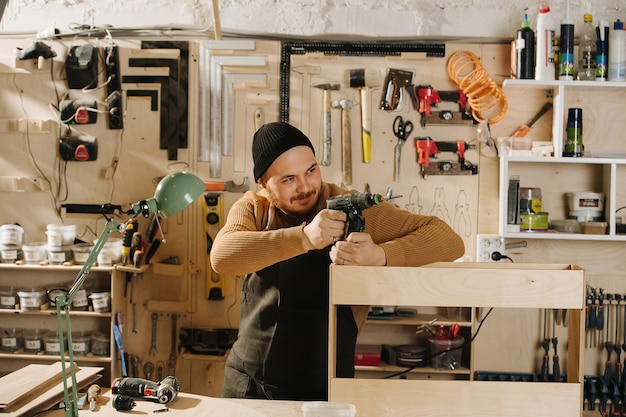 Kindlicher Tischler in einer Uhrkappe in einer großen Werkstatt. Mit einem Bohrer spielen, ihn wie eine Waffe halten und aus der Deckung zielen. Anzahl der an der Wand hängenden Werkzeuge.