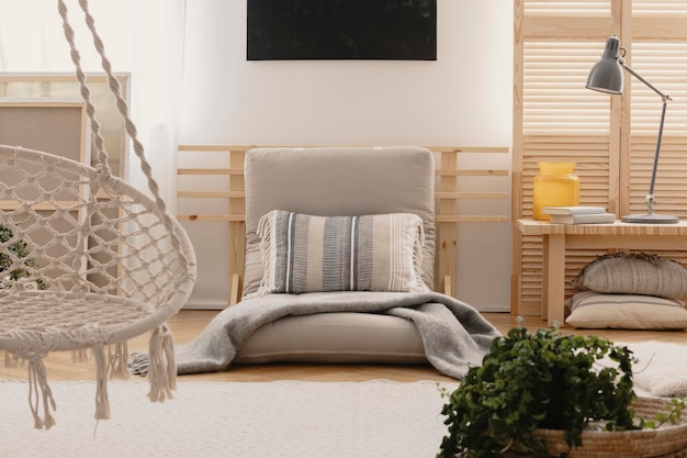 Kissen und Decke auf grauem Futon im Boho-Schlafzimmer mit Lampe und Hängesessel Echtes Foto