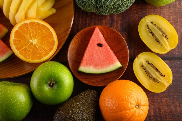 Foto kiwi de sandía naranja y piña cortado en rodajas con manzanas y brócoli en un plato de madera y una mesa de madera