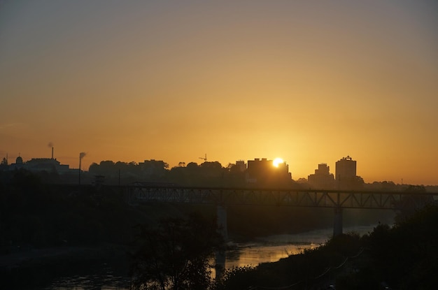 Klarer Himmel über dem Fluss und der Eisenbahnbrücke bei Sonnenaufgang. Smog über der Stadt wegen einer Fabrik mit rauchenden Pfeifen. Schutz der Umwelt vor schädlichen Emissionen