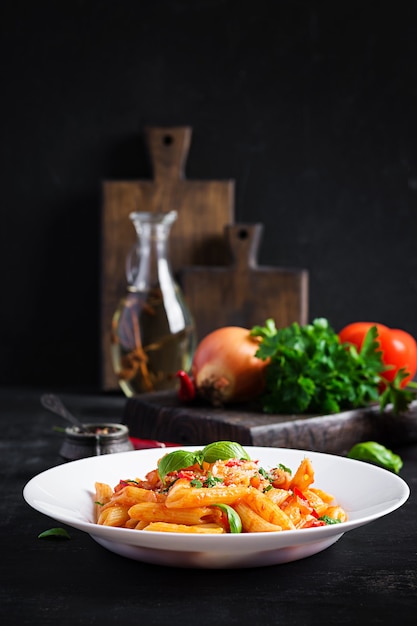 Klassische italienische Pasta Penne alla Arrabiata mit Basilikum und frisch geriebenem Parmesankäse auf dunklem Tisch. Penne-Nudeln mit Chilisauce Arrabbiata.