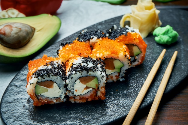 Klassische japanische Sushi-Rollen mit Lachs, Avocado, Frischkäse und Kaviar. Kalifornien rollen auf einem schwarzen Teller mit Stäbchen. Japanische Küche. Gesunde Meeresfrüchte