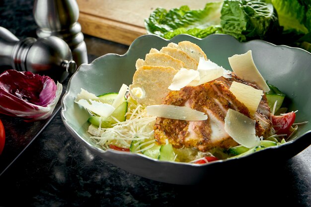 Klassischer Caesar-Salat mit Croutons, Parmesan, gegrilltem Hähnchen, serviert in einer grünen Schüssel auf einem dunklen Marmortisch. Restaurant Essen.