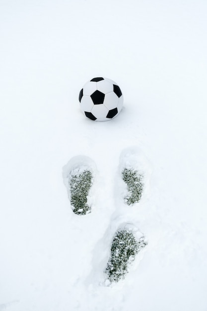 Klassischer Fußball auf einem schneebedeckten Sportplatz und Fußabdrücke im Schnee. Kopierraum, vertikale Ausrichtung