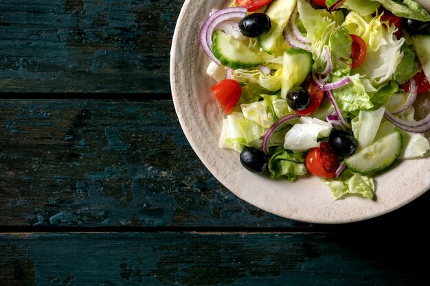 Klassischer Gemüsesalat mit Tomaten, Gurken, Zwiebeln, Blattsalaten und schwarzen Oliven in weißer Keramikplatte. Dunkler Holztisch. Flache Lage, Kopierraum