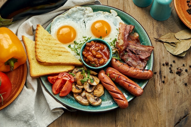 Klassisches englisches Frühstück: Toast, geräucherte Würstchen, Speck, Spiegeleier, Bohnen und gebratene Toasts auf einem blauen Teller. Draufsicht, horizontal. Holzoberfläche