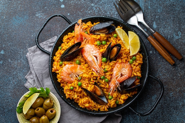 Klassisches Gericht aus Spanien, Meeresfrüchte-Paella in traditioneller Pfanne auf rustikaler blauer Betonhintergrund-Draufsicht. Spanische Paella mit Garnelen, Klammern, Muscheln, grünen Erbsen und frischen Zitronenspalten von oben