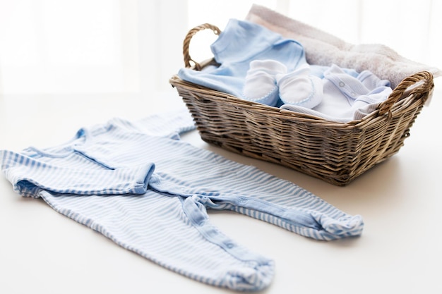 Kleidung, Babyalter, Mutterschaft und Objektkonzept - Nahaufnahme von weißen Babyschuhen mit Kleiderhaufen und Handtuch für neugeborene Jungen im Korb auf dem Tisch