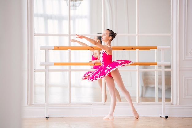 Kleine Ballerina in einem leuchtend rosa Tutu ist mit einer Ballettstange vor einem Spiegel in einem schönen weißen großen Saal beschäftigt