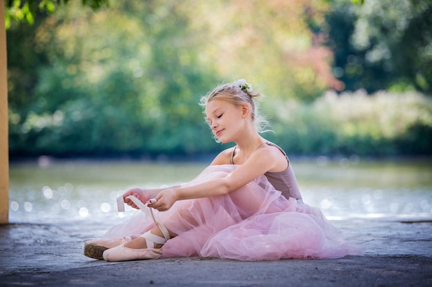 kleine ballerina in pink in einem langen ballettrock sitzt auf dem boden und bindet spitzenbänder