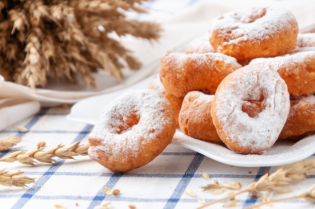 Kleine Donuts mit Puderzucker bestreut. Auf einem weißen Teller. Im Hintergrund Weizenähren und Körner.