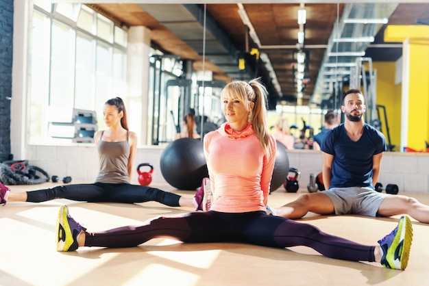 Kleine Gruppe von Menschen, die mit gespreizten Beinen auf dem Boden des Fitnessraums sitzen.