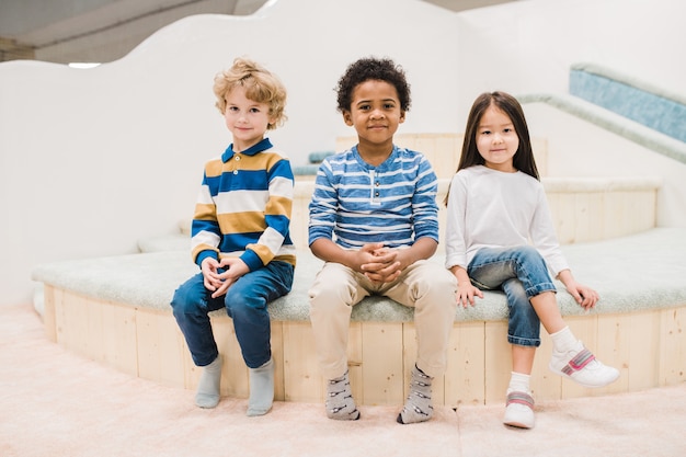 Foto kleine interkulturelle gruppe von niedlichen kleinen kindern, die vor der kamera im modernen spielzentrum für kinder sitzen