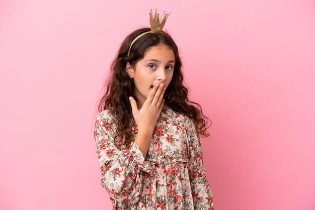 Kleine kaukasische Prinzessin mit Krone isoliert auf rosa Hintergrund, glücklich und lächelnd, den Mund mit der Hand bedeckend