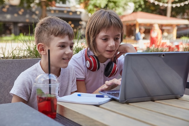 Kleine kaukasische Schulkinder sitzen im Freien auf dem Bildschirm des Laptops und haben Spaß