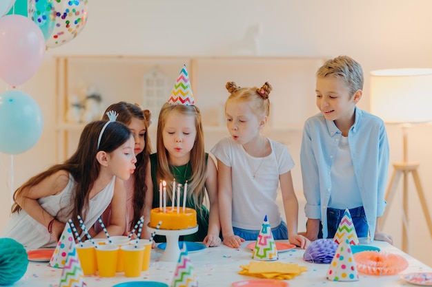 Kleine Kinder feiern Geburtstagsparty, blasen Kerzen auf Kuchen, versammeln sich am festlichen Tisch, haben gute Laune, verbringen gerne Zeit miteinander, machen Wunsch, tragen Partyhüte, posieren drinnen mit aufgeblasenen Luftballons