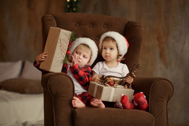 Kleine Kinder mit Weihnachtsmannmützen sitzen auf einem gemütlichen Stuhl im Zimmer
