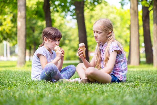 Kleine lächelnde Mädchenfreundinnen, die auf dem Rasen sitzen und Eiscreme essen. Kinderferien