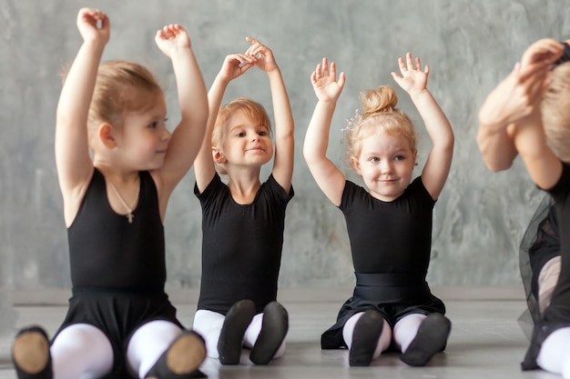 Kleine Mädchen Ballerina in schwarzen Kleidern, Strumpfhosen und Spitzenschuhen sitzen auf dem Boden und halten die Hände über dem Kopf zum Aufwärmen, bevor sie Ballett in einem dunklen Tanzstudio unterrichten