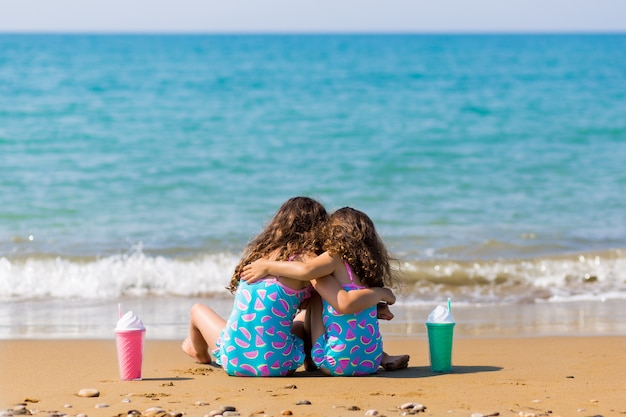 Kleine Mädchen lehnen sich im Sand zurück und kuscheln mit Cocktails. Familienurlaub Konzept. glückliche Schwestern