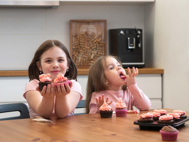 Kleine Mädchen mit Cupcakes, von denen eines Süßigkeiten hält, das andere lächelt und sich die Finger leckt