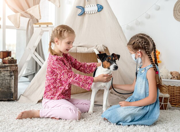 Kleine Mädchen mit Phonendoskop hörendem Hund wie Arzt, der auf Boden im Spielzimmer sitzt