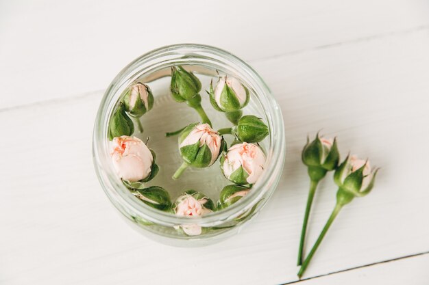 kleine rosa Rosen mit Knospen im Glasgefäß auf weißem Holzbretthintergrund, Draufsicht