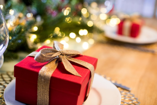 Kleine rote Geschenkboxen mit goldenem Band auf weißen Tellern als Teil der Weihnachtstischdekoration Überraschung für jeden Gast Tanne Zweig mit Lichtern auf Hintergrund blured Selektiver Fokus