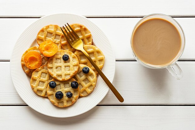 Kleine runde Waffeln mit Früchten und Kaffee auf weißem Holzhintergrund, flach gelegt