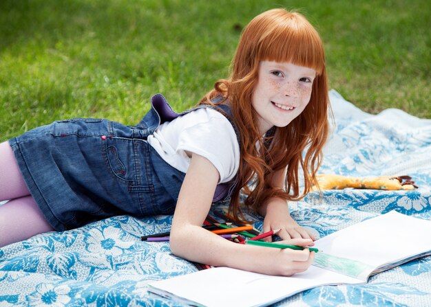 Kleine schöne lächelnde Mädchenzeichnung mit Bleistiften auf dem Plaid auf dem Gras