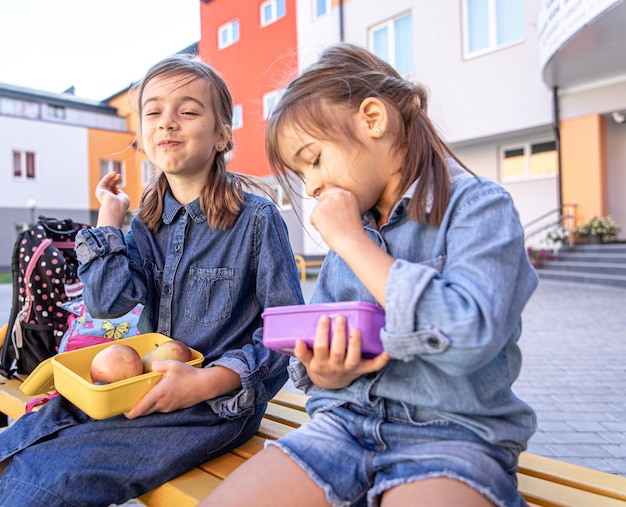 Kleine Schulmädchen sitzen auf der Bank im Schulhof und essen aus Brotdosen.