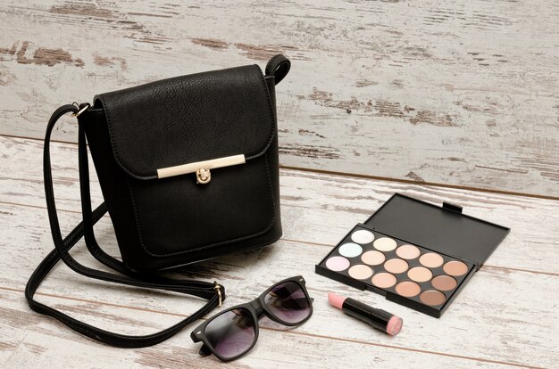 Kleine schwarze Damenhandtasche, Sonnenbrille, Lidschattenpalette und Lippenstift auf Holz. Mode