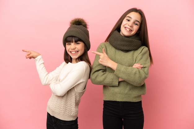 Kleine Schwestern, die eine Winterkleidung tragen, lokalisiert auf rosa Hintergrund, der Finger zur Seite in seitlicher Position zeigt