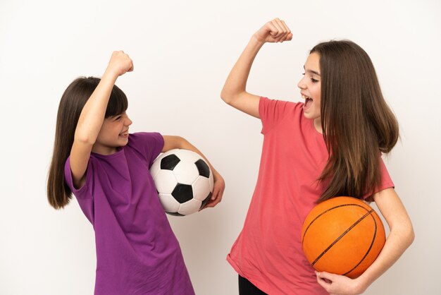 Kleine Schwestern, die Fußball und Basketball spielen, isoliert auf weißem Hintergrund, die einen Sieg in Siegerposition feiern