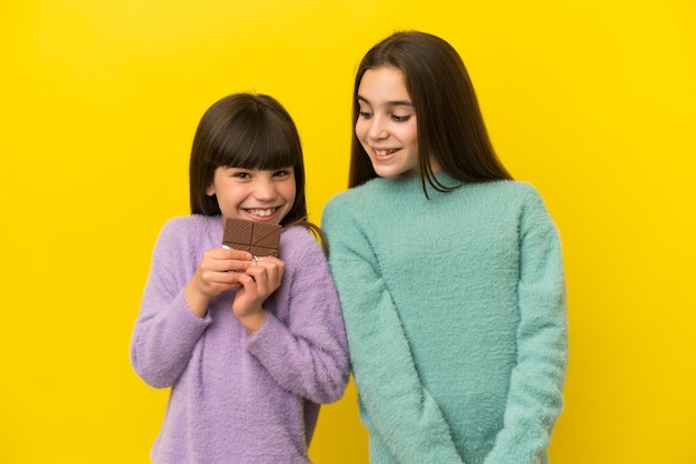 Kleine Schwestern lokalisiert auf gelbem Hintergrund, die eine Schokoladentafel nehmen und glücklich