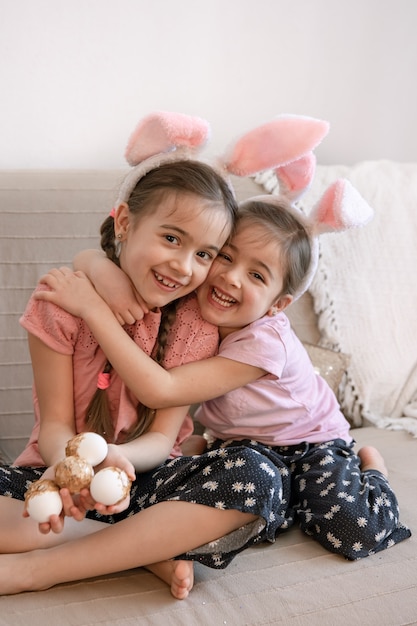 Kleine Schwestern mit Hasenohren und Ostereiern umarmen sich sitzend auf der Couch. Ostern Fotosession Konzept.