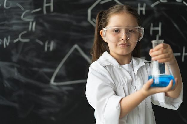 Kleine Studentin mit Brille in einem Laborkittel steht in der Nähe der Tafel