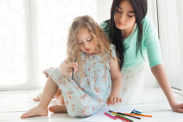 Kleine süße Tochter und ihre Mutter zeichnen mit bunten Stiften und haben Spaß zusammen. Hübsches Kind und Mutter spielen drinnen. Glückliche Familie, die Zeit durch Zeichnen verbringt.