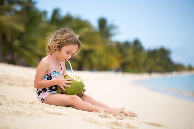 Kleine Vorschulkindfrau, die Kokosnusssaft am Ozeanstrand trinkt