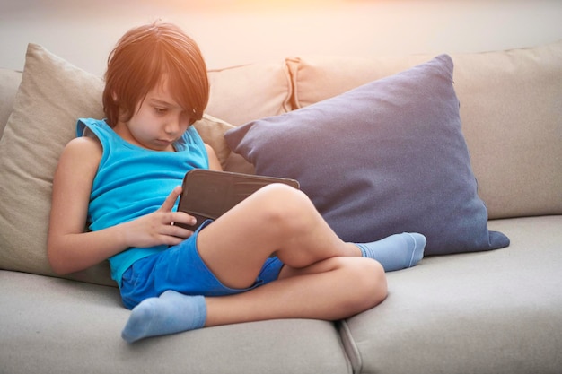 Kleiner arabischer Junge sitzt auf Sofa und spielt Spiel auf digitalem Tablet. Porträt eines jungen arabischen Kindes, das sich zu Hause Zeichentrickfilme auf dem Laptop ansieht. Moderne E-Learning-Technologie für Kinder- und Online-Bildung.