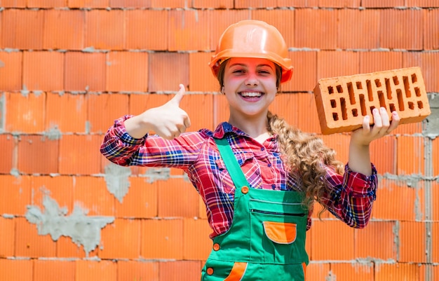 Kleiner Arbeiter im Helmbau aus Backstein Architektur Baukind Kind in Uniform, das um die Ziegelmauer herum arbeitet Tag der Arbeit Konzept Kleines Mädchen Maurer lernt, wie man den Daumen nach oben baut